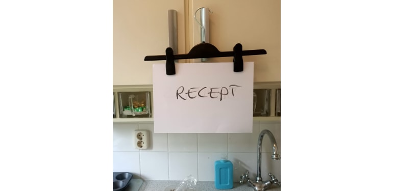 Een broekenhanger met een blad papier waarop recept geschreven staat. Deze hangt aan een keukenkastje. 