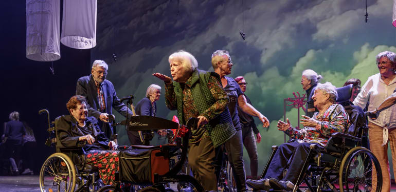 Groep oudere acteurs op het toneel tijdens een voorstelling. Een aantal zit in een rolstoel.